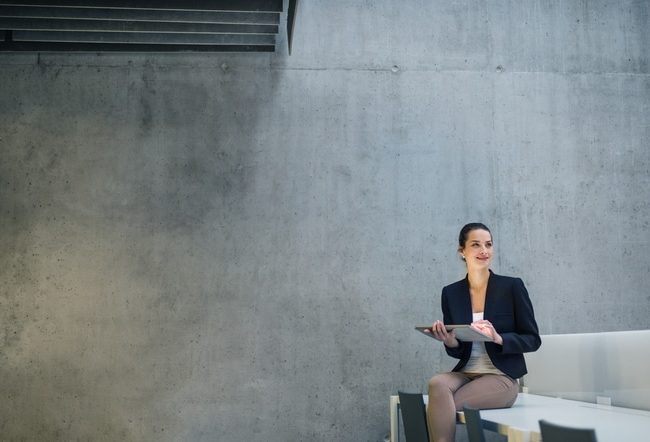 Young business woman with tablet sitting on desk against concrete wall in office. Copy space.